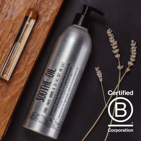 A 400ml aluminium bottle with a black pump lying diagonally on a grey slate, next to 3 sprigs of dried lavender. To the left of the bottle is a wooden board with a clear glass testube of oil lyig on it with a cork stopper.