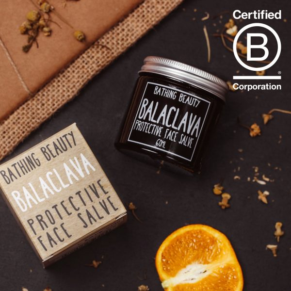 An amber glass jar of balaclava face salve lying on a gray slate. Next to it is a cut mandarin, and a sprinkling of chamomile flowers. to its left is the Balaclava product box. Proudly displaying the B Corp Logo in the top right corner