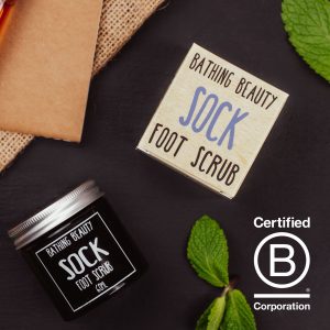 An amber jar of Sock Foot Scrub with an aluminium lid lying on a Grey Slate. Next to it is some sprigs of mint. a B Corp Logo, and the product box