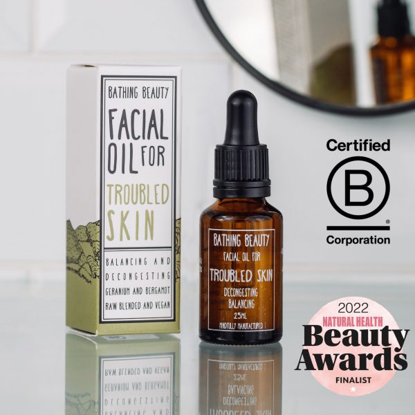 An amber glass bottle with black pipette on a glass shelf. To the left is a green and white box. to the right a round mirror in a black frame and two logos. Natural health and beauty finalist and B Corp Corporation
