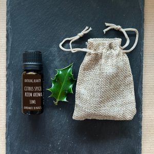 Amber glass bottle of sweet orange and cinnamon essential oil lying on a dark grey slate next to a holly sprig and a mini draw string Jute bag
